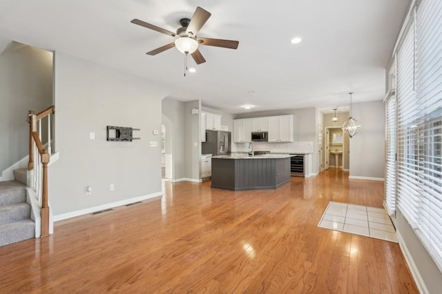 unfurnished living room with light wood finished floors, beverage cooler, visible vents, stairway, and ceiling fan with notable chandelier