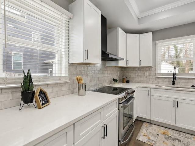 kitchen featuring a tray ceiling, stainless steel electric stove, a sink, wall chimney range hood, and backsplash