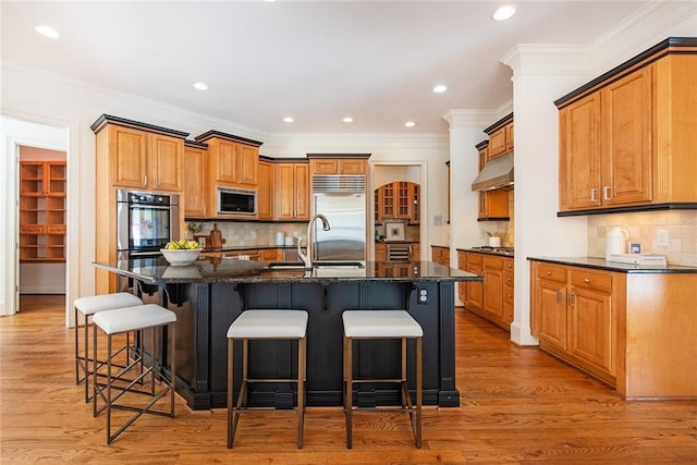 kitchen with an island with sink, built in appliances, dark stone countertops, and light hardwood / wood-style flooring