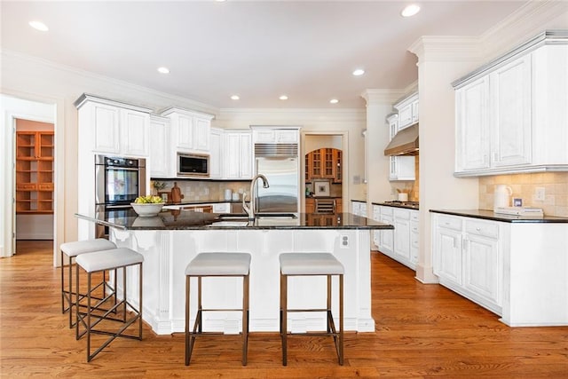 kitchen featuring built in appliances, tasteful backsplash, a kitchen island with sink, white cabinetry, and light wood-type flooring