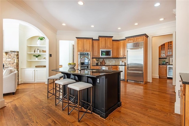 kitchen featuring a kitchen bar, dark stone counters, built in appliances, hardwood / wood-style flooring, and an island with sink