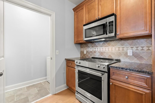 kitchen with stainless steel appliances, backsplash, brown cabinetry, dark stone counters, and baseboards