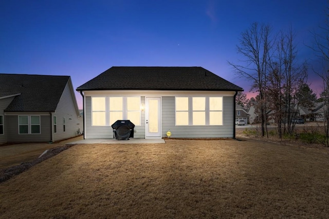 back of property at dusk featuring roof with shingles, a lawn, and a patio