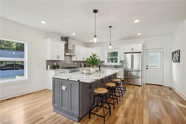 kitchen with white cabinetry, hanging light fixtures, an island with sink, stainless steel appliances, and wall chimney range hood