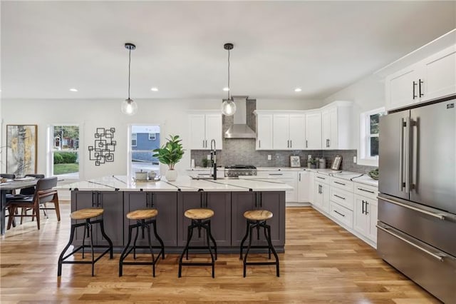 kitchen with pendant lighting, a kitchen island with sink, high quality fridge, white cabinets, and wall chimney exhaust hood