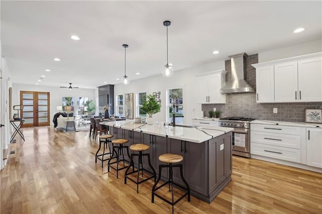 kitchen featuring high end range, hanging light fixtures, a center island with sink, wall chimney range hood, and white cabinets