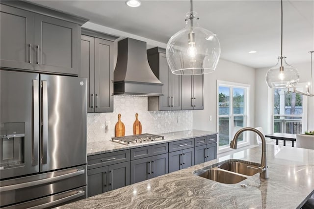 kitchen with gray cabinets, a sink, appliances with stainless steel finishes, wall chimney range hood, and light stone countertops