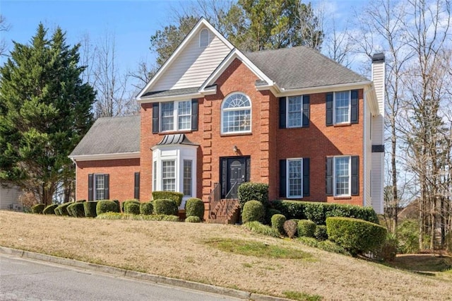 colonial inspired home featuring brick siding, a chimney, a shingled roof, and a front lawn