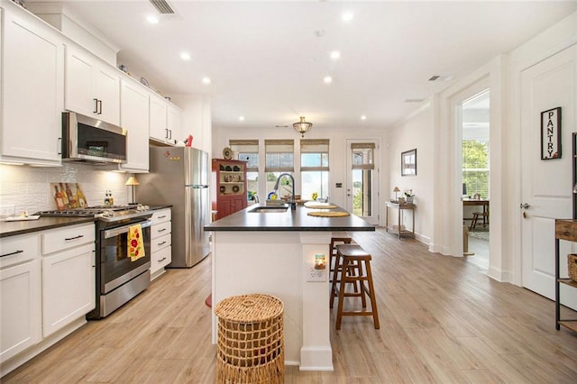 kitchen with dark countertops, appliances with stainless steel finishes, a center island with sink, and light wood-type flooring
