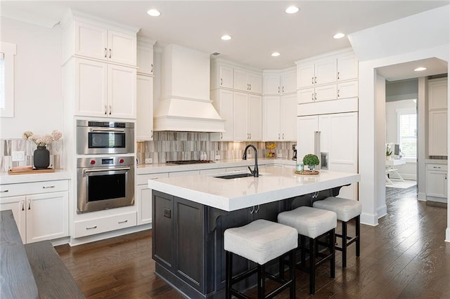 kitchen featuring white cabinets, a sink, premium range hood, tasteful backsplash, and dark wood-style floors