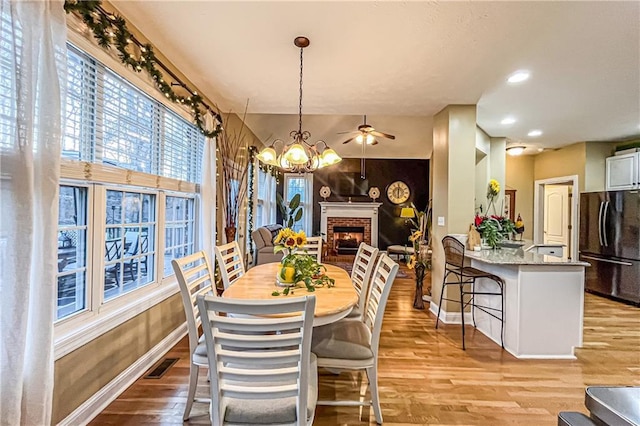 dining space with light wood-type flooring, ceiling fan with notable chandelier, and a brick fireplace