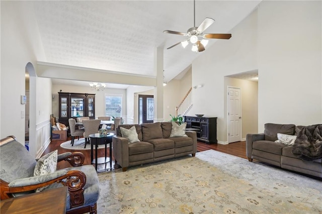 living room featuring dark wood finished floors, stairway, ceiling fan with notable chandelier, arched walkways, and high vaulted ceiling