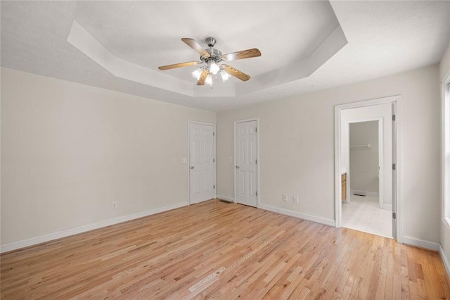 unfurnished bedroom with a ceiling fan, light wood-type flooring, a raised ceiling, baseboards, and ensuite bathroom