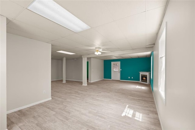 below grade area featuring ceiling fan, a warm lit fireplace, baseboards, a drop ceiling, and wood finished floors