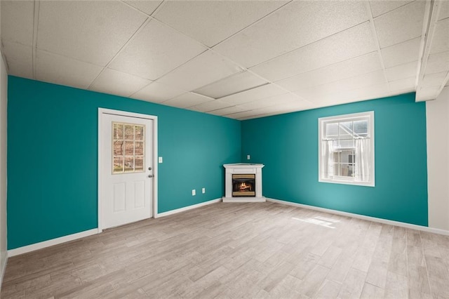 unfurnished living room featuring baseboards, a drop ceiling, a warm lit fireplace, and wood finished floors