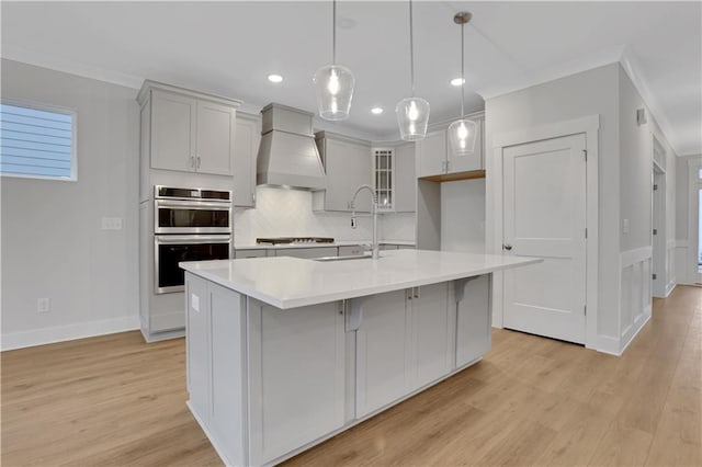 kitchen featuring pendant lighting, an island with sink, stainless steel double oven, and premium range hood