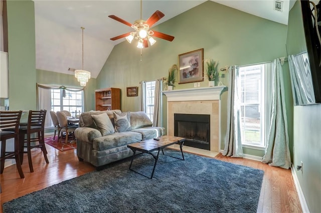 living room with ceiling fan with notable chandelier, wood finished floors, high vaulted ceiling, visible vents, and a tiled fireplace
