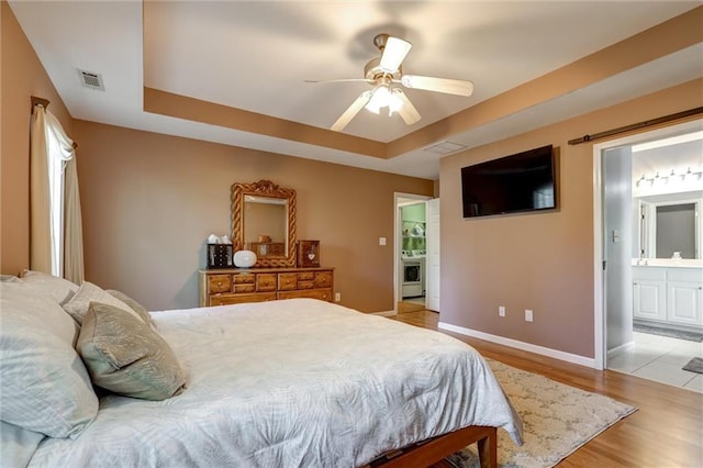 bedroom with ensuite bath, visible vents, light wood finished floors, a raised ceiling, and baseboards