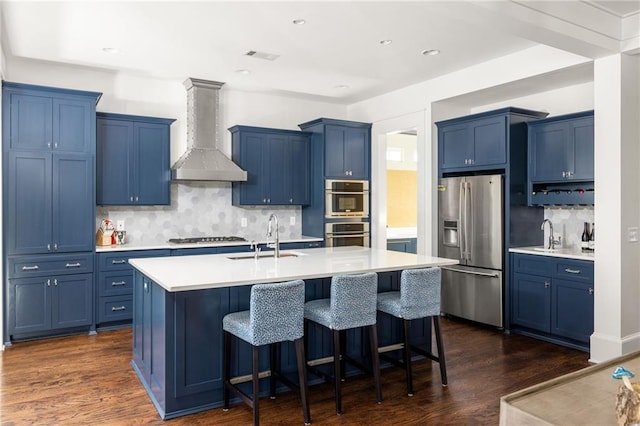 kitchen featuring wall chimney exhaust hood, sink, stainless steel appliances, and an island with sink