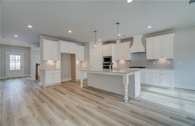 kitchen featuring premium range hood, white cabinetry, light countertops, an island with sink, and decorative light fixtures