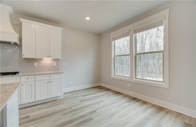 kitchen featuring baseboards, light wood-style floors, white cabinets, backsplash, and custom exhaust hood