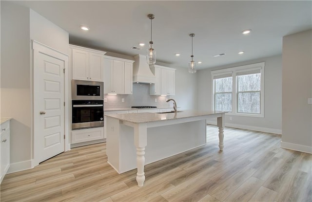 kitchen with stainless steel appliances, white cabinets, custom exhaust hood, a center island with sink, and decorative light fixtures