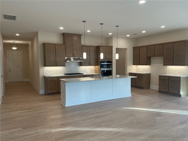 kitchen with pendant lighting, double oven, stainless steel gas stovetop, light stone counters, and a center island with sink