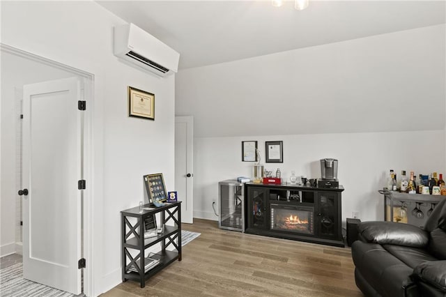 living area with a glass covered fireplace, wood finished floors, a wall unit AC, baseboards, and vaulted ceiling