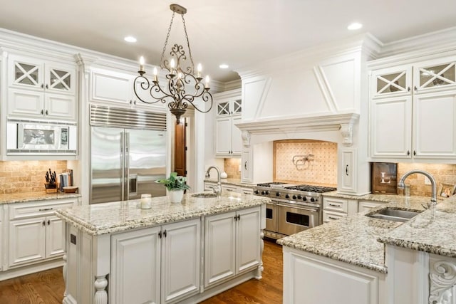 kitchen with sink, built in appliances, white cabinets, hanging light fixtures, and an island with sink