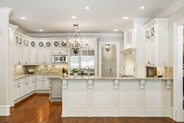 kitchen featuring light stone counters, a kitchen island with sink, built in appliances, decorative light fixtures, and white cabinets