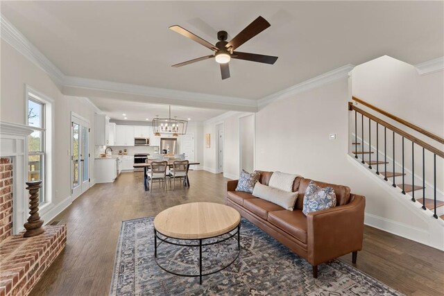 living room featuring a brick fireplace, ceiling fan with notable chandelier, crown molding, and dark hardwood / wood-style floors