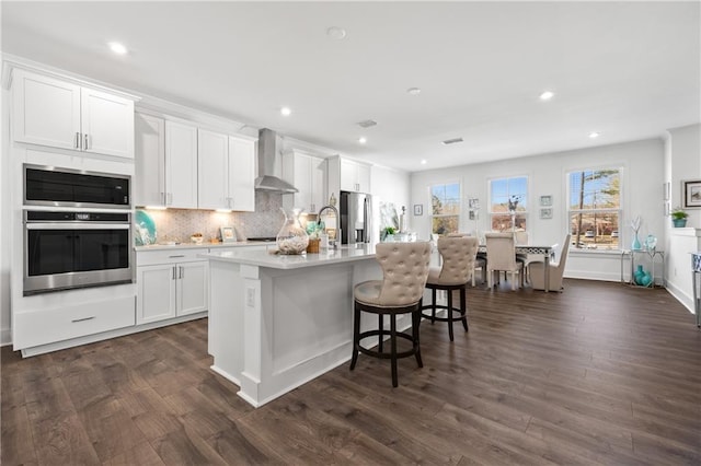 kitchen featuring a kitchen island with sink, wall chimney range hood, tasteful backsplash, stainless steel fridge with ice dispenser, and light countertops