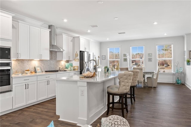 kitchen featuring dark wood finished floors, stainless steel appliances, light countertops, a kitchen breakfast bar, and wall chimney range hood