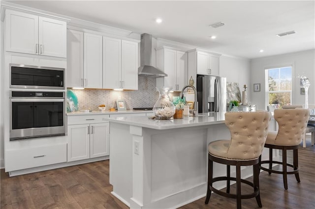 kitchen featuring an island with sink, tasteful backsplash, appliances with stainless steel finishes, wall chimney exhaust hood, and light countertops