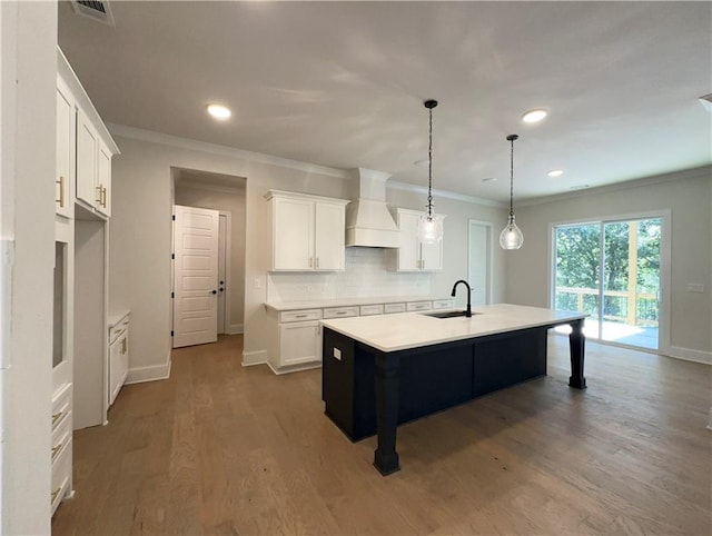 kitchen with custom range hood, a kitchen island with sink, sink, pendant lighting, and white cabinets