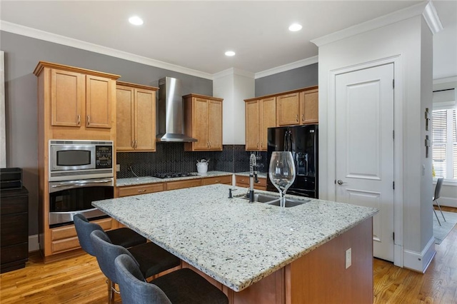 kitchen with light wood-style floors, wall chimney exhaust hood, a sink, black appliances, and backsplash