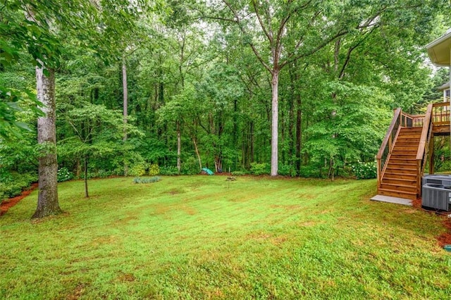 view of green lawn featuring stairs, central AC unit, a wooded view, and a wooden deck