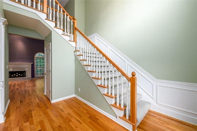 stairs with wood finished floors, a decorative wall, a towering ceiling, and a fireplace with raised hearth