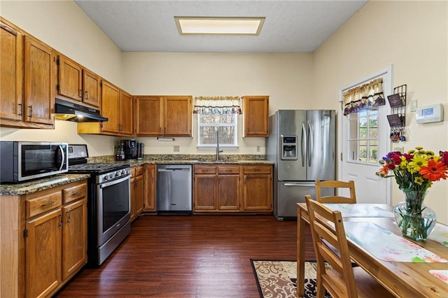 kitchen with under cabinet range hood, a sink, appliances with stainless steel finishes, brown cabinetry, and dark wood-style flooring