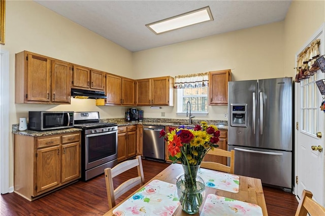 kitchen with brown cabinets, under cabinet range hood, a sink, dark wood-style floors, and stainless steel appliances