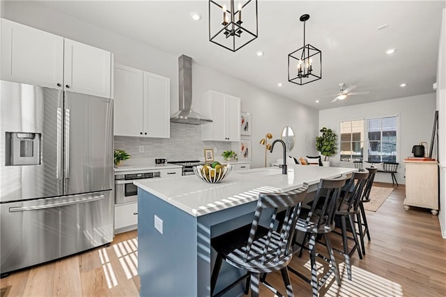 kitchen with an island with sink, a sink, wall chimney range hood, appliances with stainless steel finishes, and light countertops