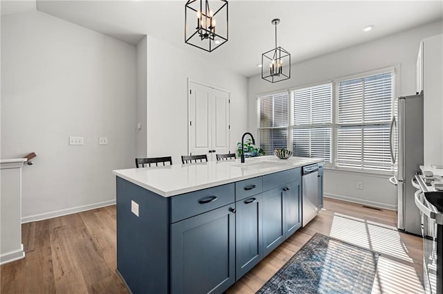 kitchen with an island with sink, a sink, stainless steel appliances, light wood-type flooring, and a chandelier