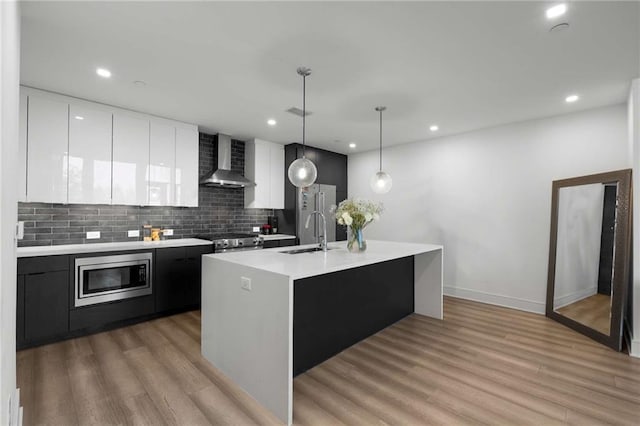 kitchen with appliances with stainless steel finishes, light countertops, light wood finished floors, dark cabinets, and wall chimney exhaust hood