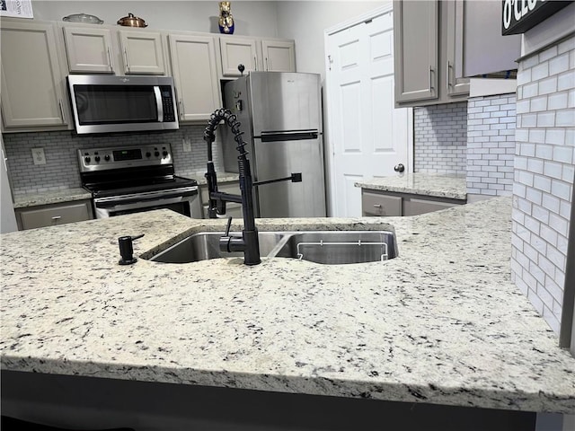 kitchen with appliances with stainless steel finishes, a sink, backsplash, gray cabinets, and light stone countertops