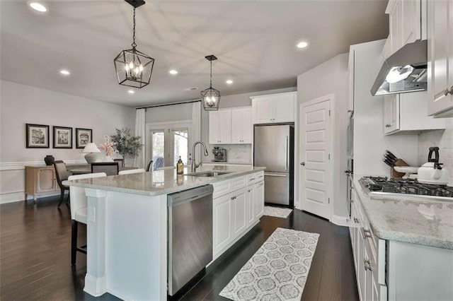 kitchen with wall chimney exhaust hood, white cabinetry, appliances with stainless steel finishes, pendant lighting, and a kitchen island with sink