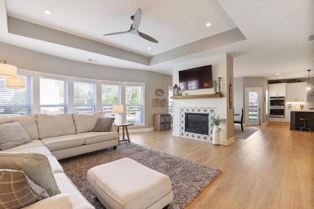 living area featuring light wood-style floors, a fireplace, a tray ceiling, a ceiling fan, and baseboards