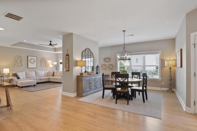 dining room with ceiling fan with notable chandelier, a tray ceiling, light wood-type flooring, visible vents, and baseboards