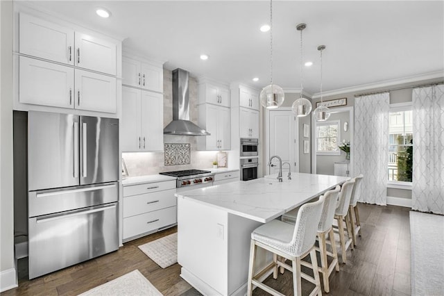 kitchen with white cabinetry, a kitchen island with sink, decorative light fixtures, appliances with stainless steel finishes, and wall chimney range hood