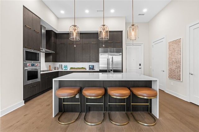 kitchen featuring built in appliances, light hardwood / wood-style floors, dark brown cabinets, and a center island with sink