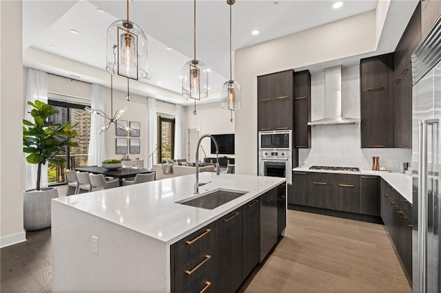 kitchen featuring sink, hanging light fixtures, light hardwood / wood-style flooring, a center island with sink, and wall chimney range hood